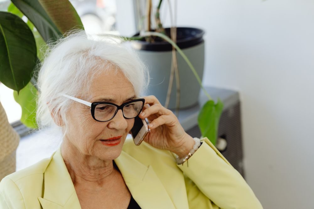 Ältere Frau mit Brille telefoniert zu Hause neben Zimmerpflanzen